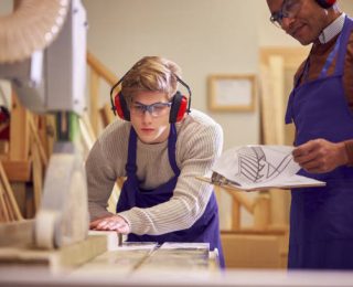 Tutor With Male Carpentry Student In Workshop Studying For Apprenticeship At College Using Bench Saw